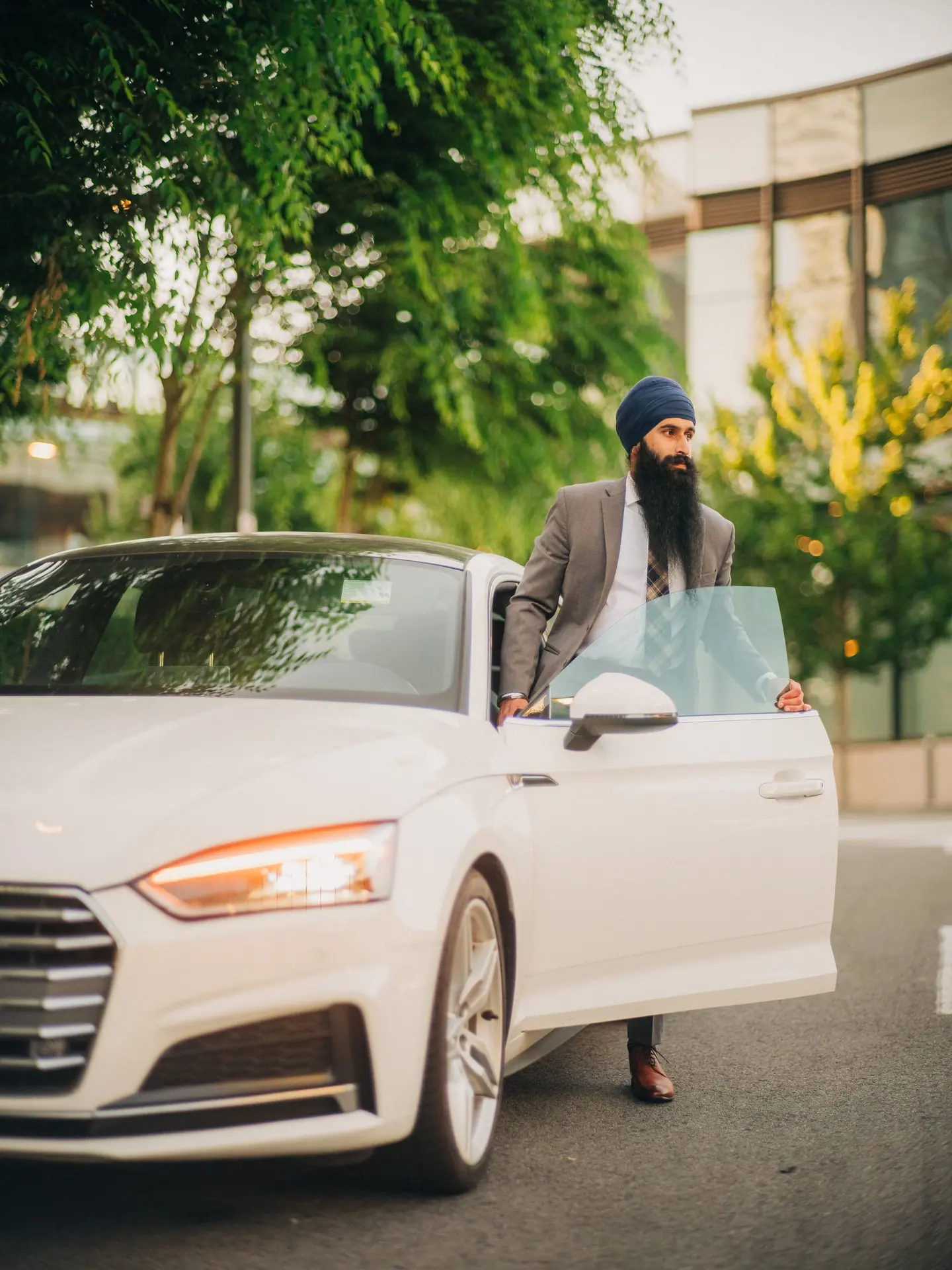 A man in a grey suit and navy turban standing at the open door of a white car on a tree lined city street.