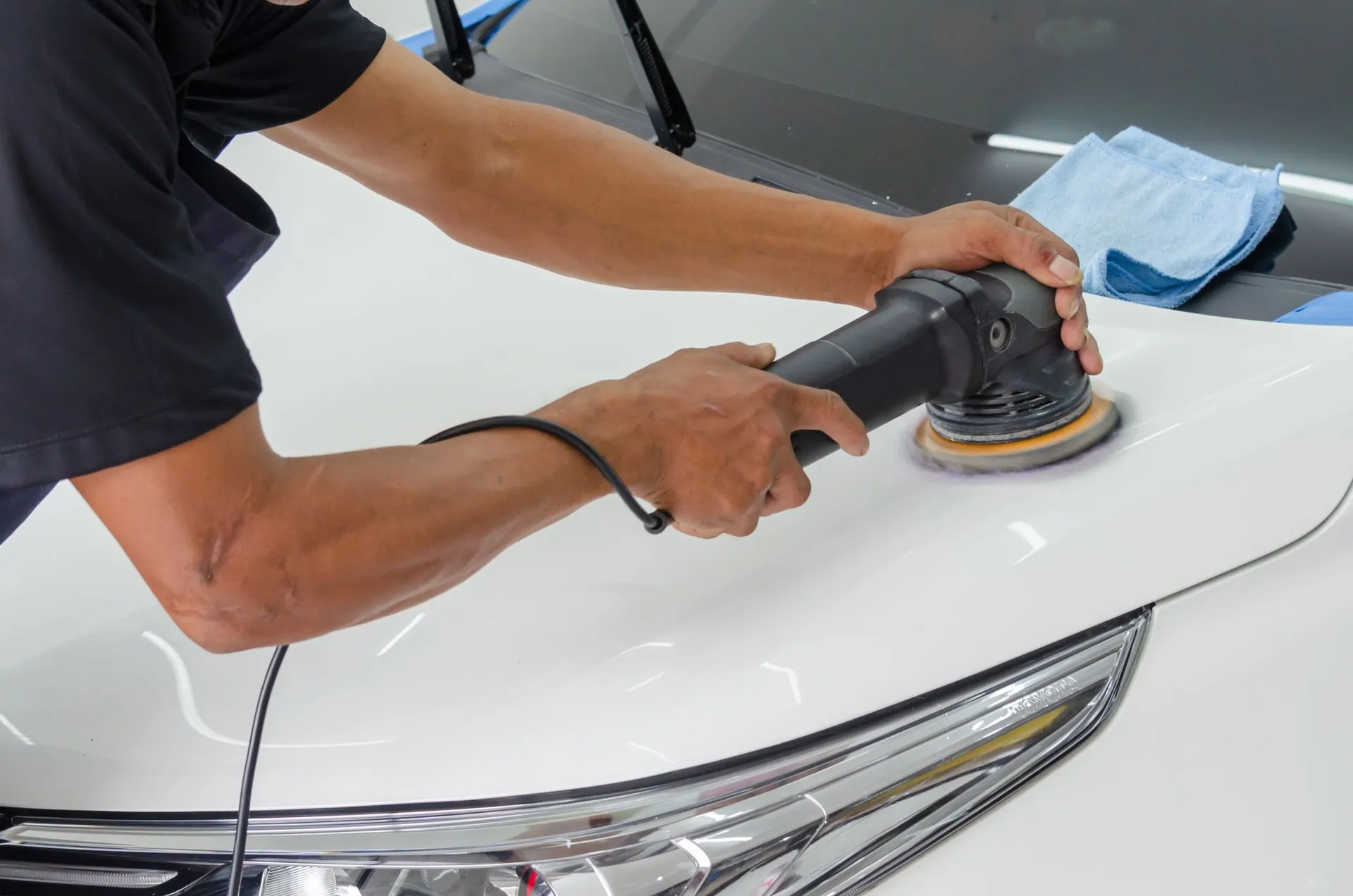 A man in a dark polo shirt guiding a dual action polisher with a cream foam pad across the white bonnet of a car, a blue microfibre cloth folded behind it.