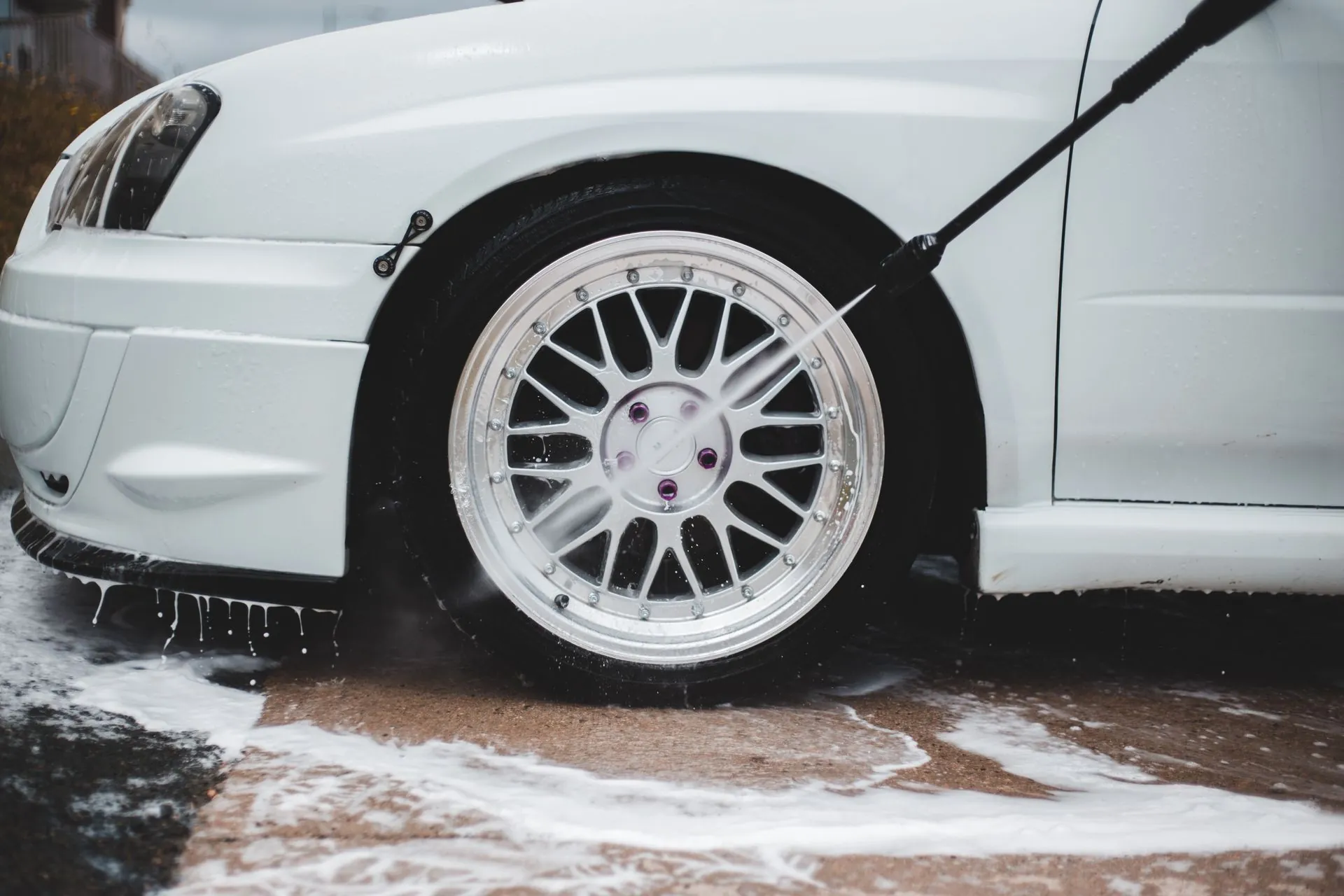 A pressure lance rinsing foam off the front wheel and arch of a white car.