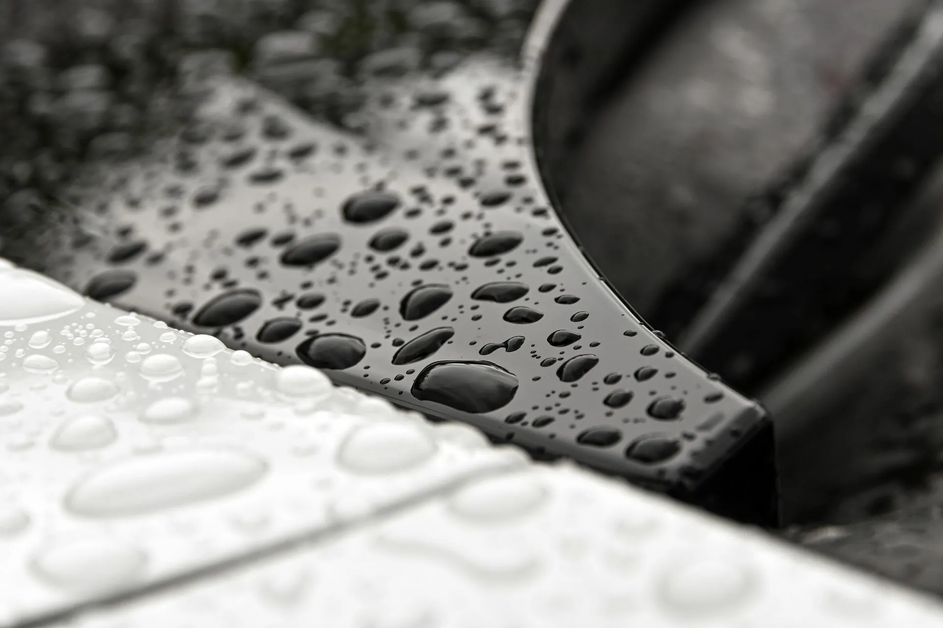 Close up of rain droplets standing in rounded beads across a black car panel beside a white one, shot from a low angle with the background blurred.